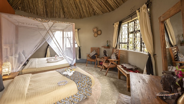 A room with a thatched roof, two beds with mosquito nets, wooden furniture, and a large window with curtains.