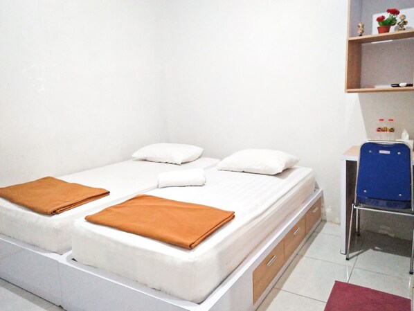 A single-bed room with a white mattress, two pillows, and two folded blankets. There is a small shelf with decorative items and a blue chair. - De Cengger Guest House (Malang)