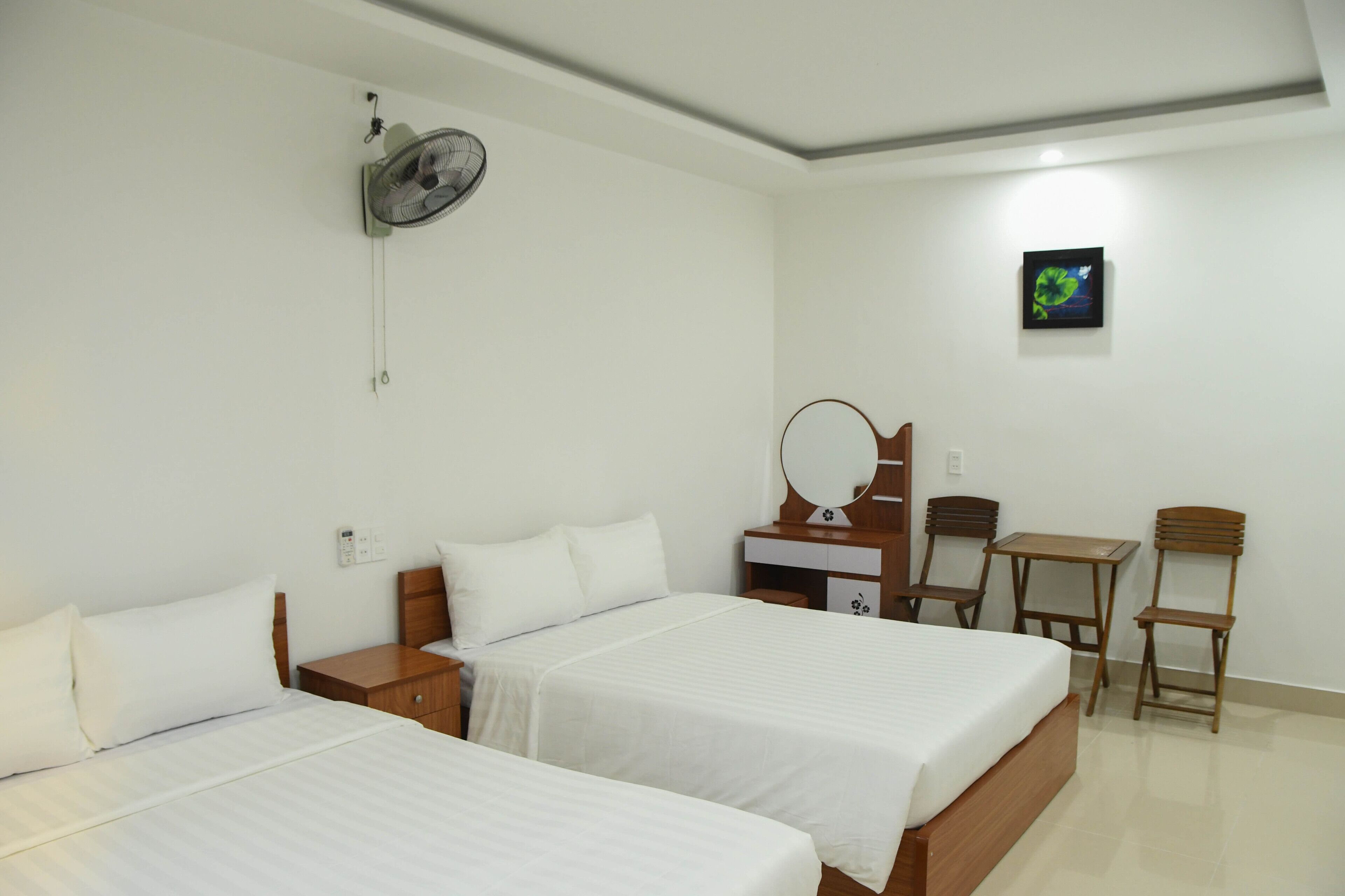 A hotel room with two beds, a desk, a chair, a small table, a mirror, and a wall-mounted fan.