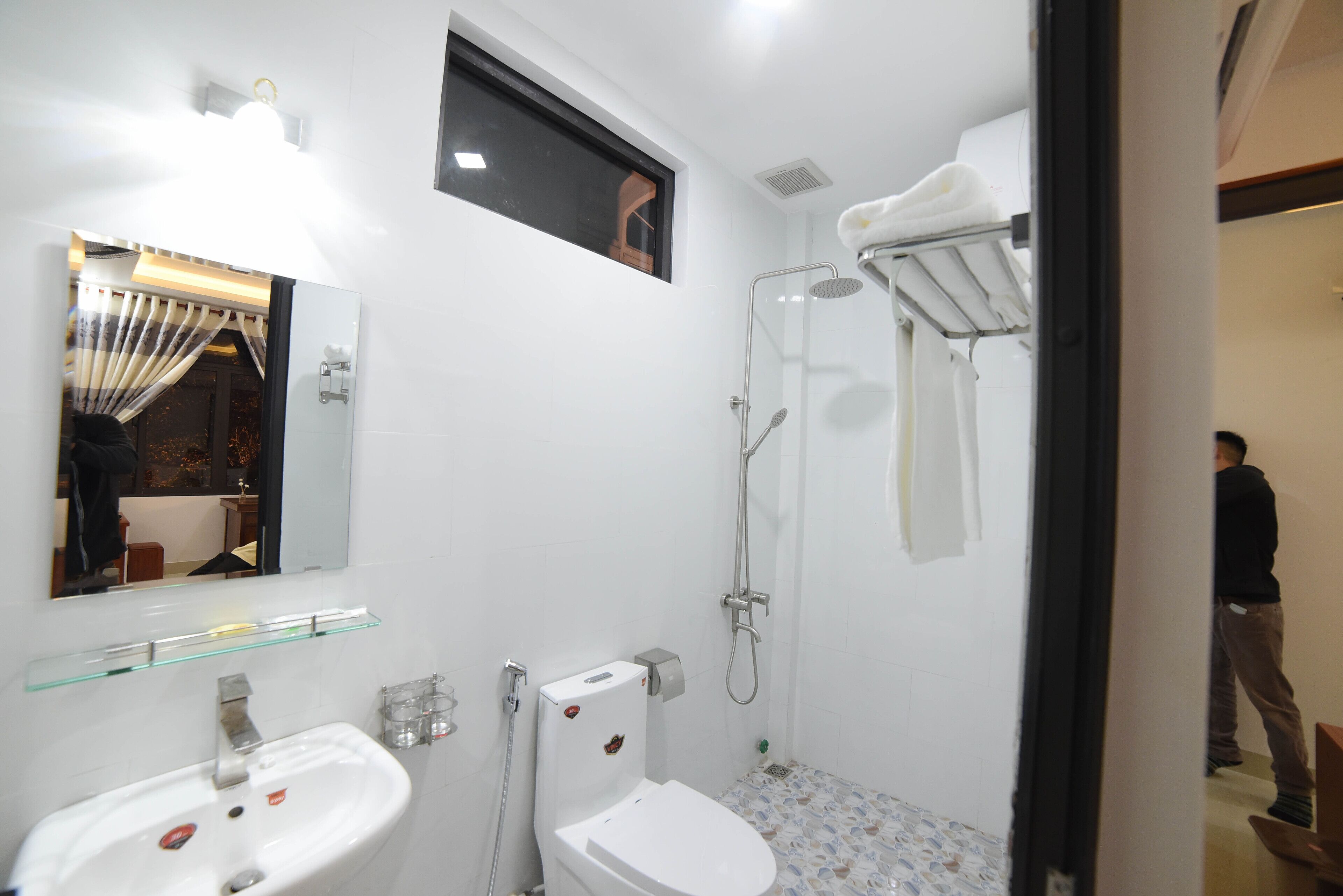 Uma casa de banho moderna com uma prateleira de vidro sobre a pia, uma sanita com dispensador de papel e um chuveiro com barra para toalhas.