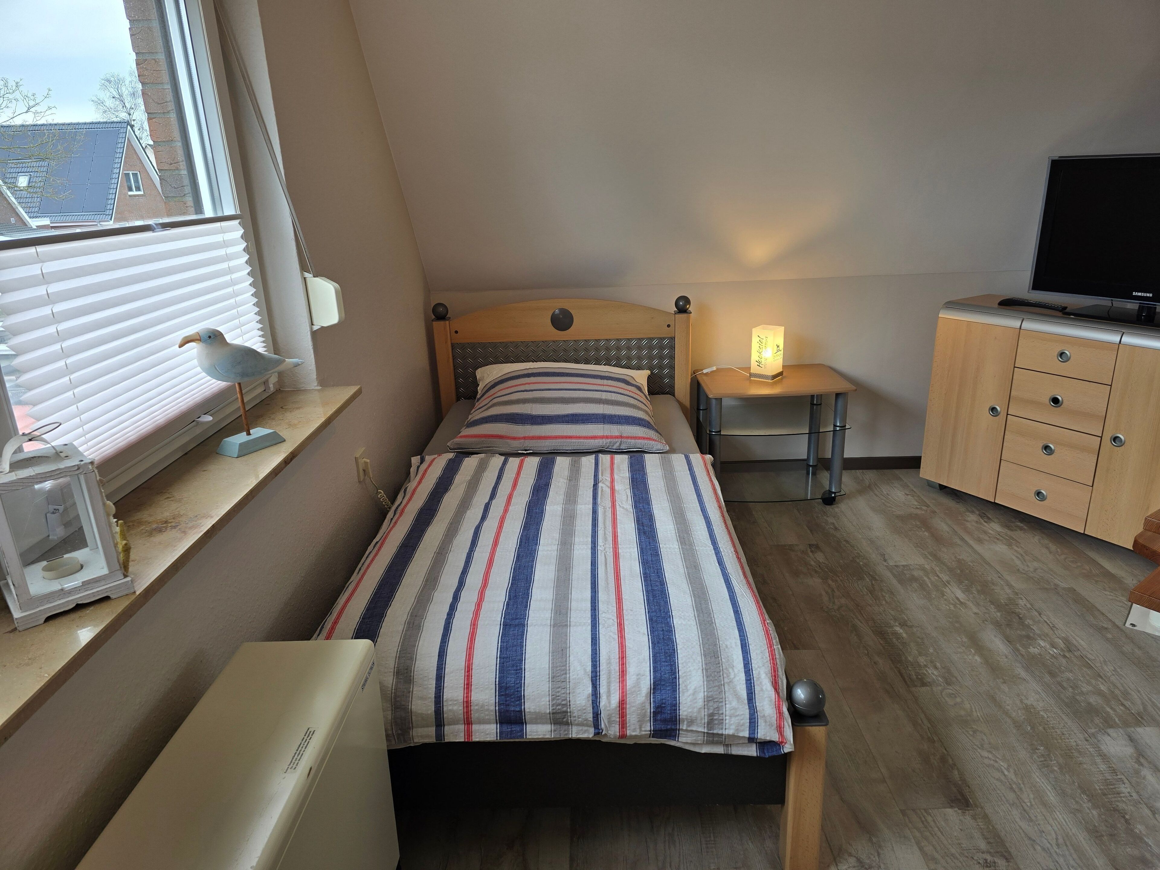 A small, neatly arranged bedroom with a single bed, a bedside table with a lamp, a TV on a stand, and a window with blinds.