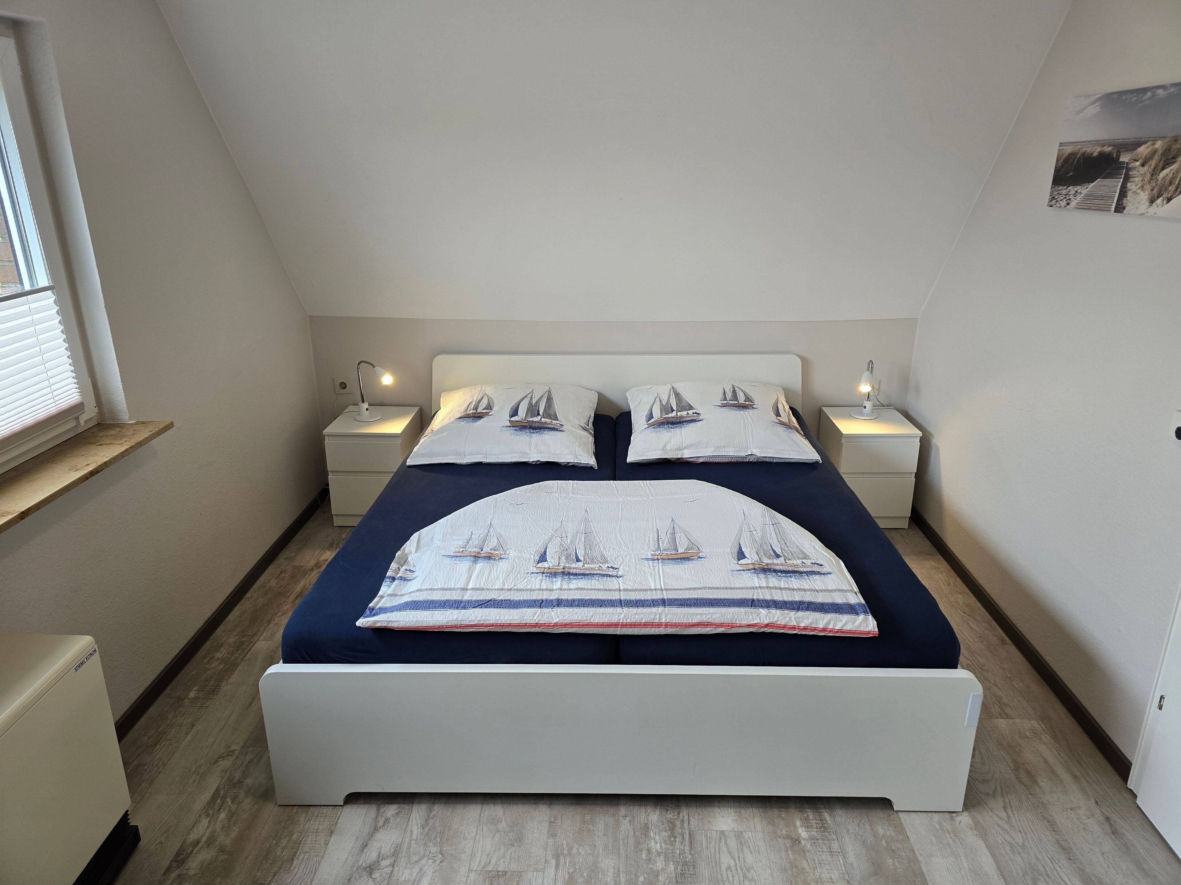 Un dormitorio con una cama, dos mesitas de noche, una mesilla auxiliar, una ventana y un cuadro colgado en la pared.