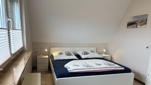 A bedroom with a bed, two bedside tables, a nightstand, a window with blinds, and a wall-mounted picture. - House Wattkieker, Germany (Wangerland)