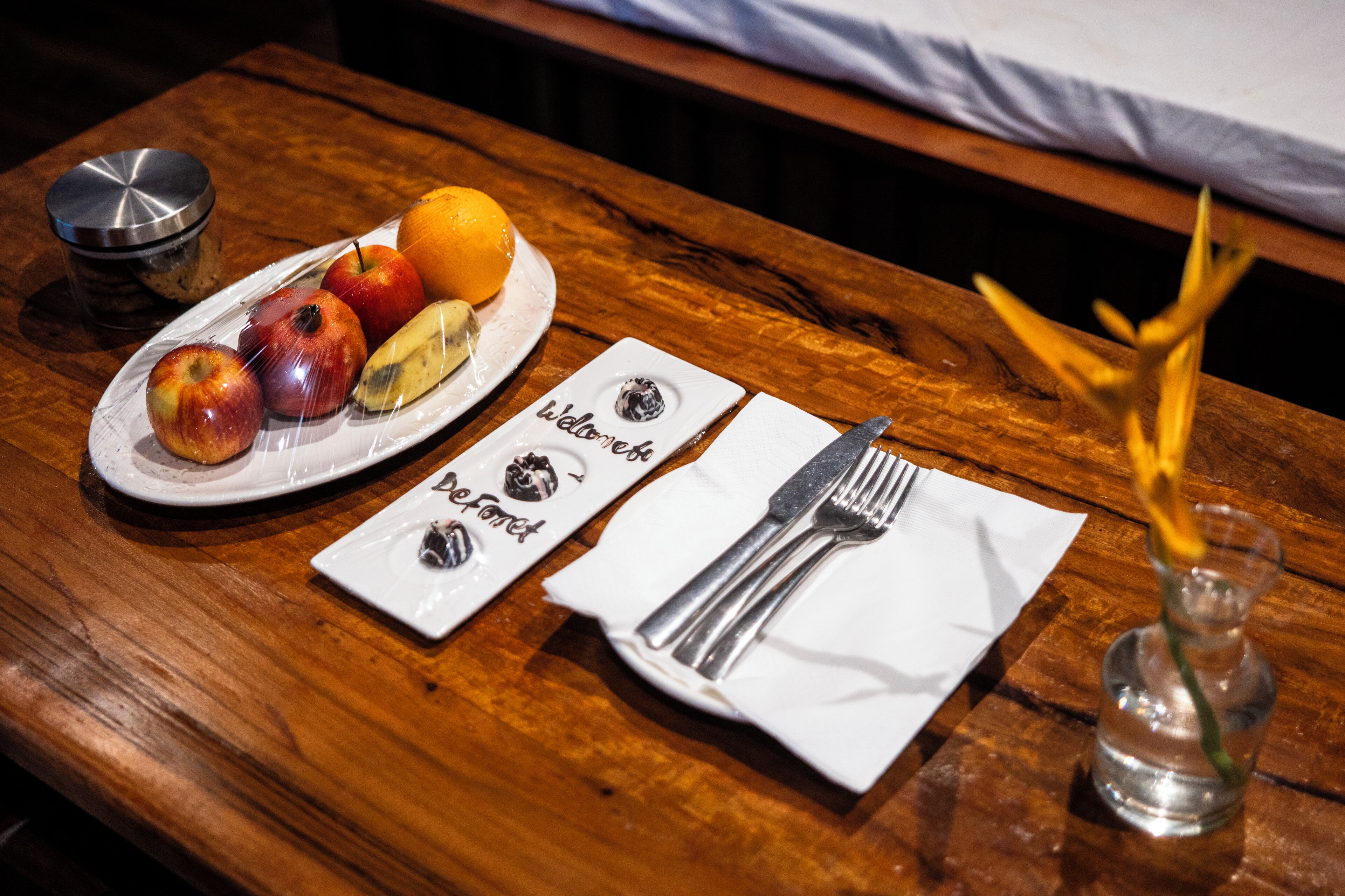 A wooden table with a plate of fruit, a glass of water, and a napkin with a fork and knife.
