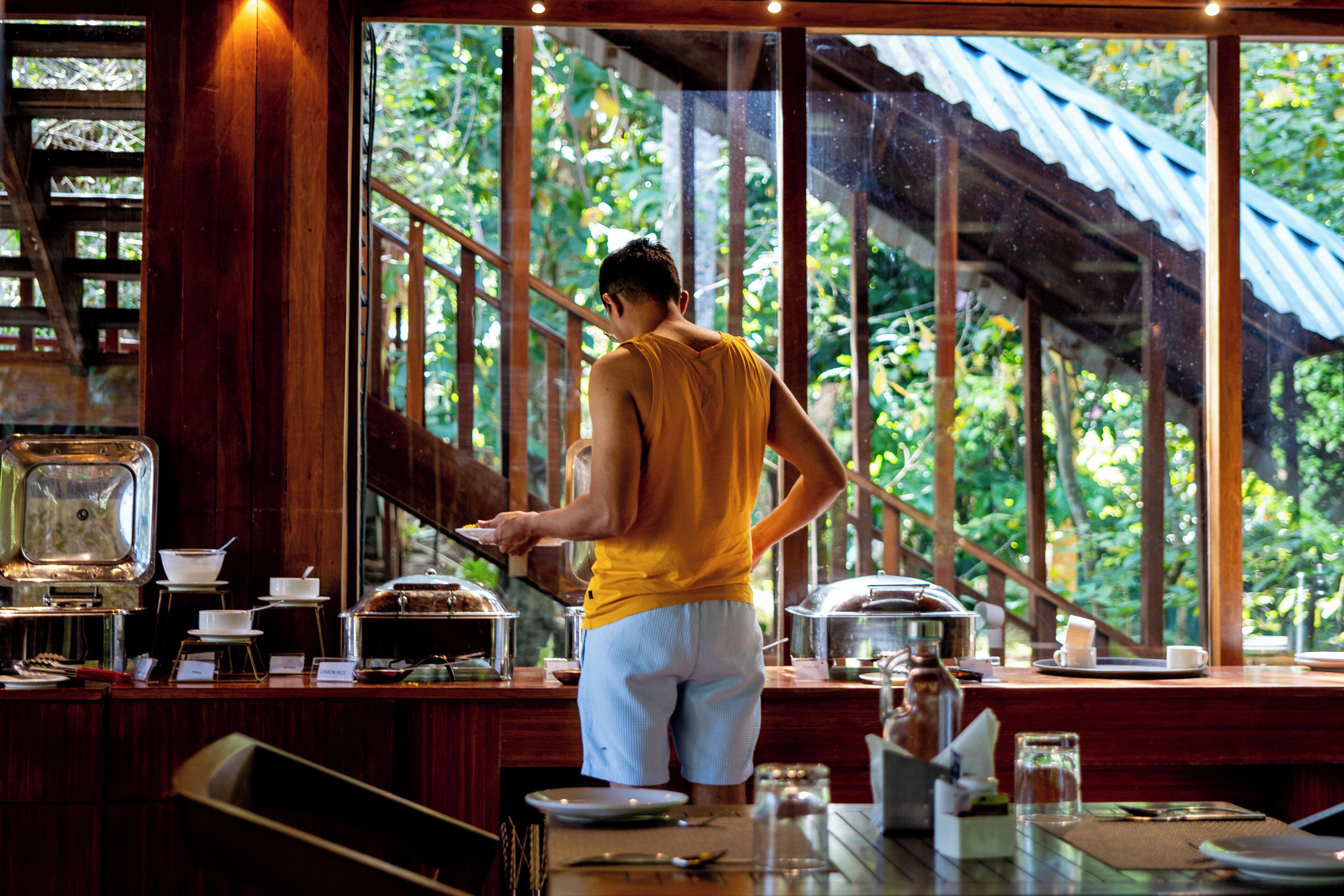 A person serving food at a buffet with a view of greenery outside.