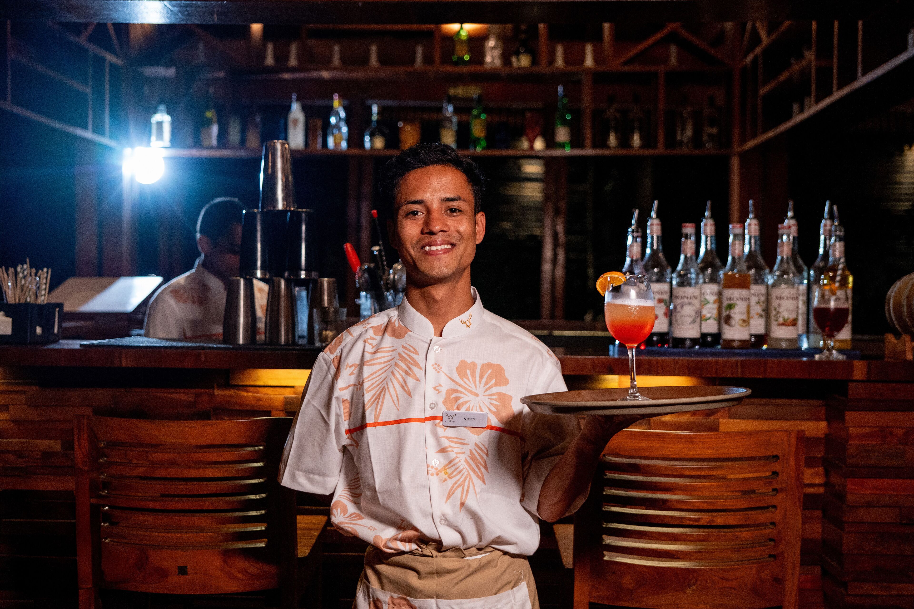 A bar setting with wooden furniture, a bartender holding a tray, and a variety of bottles in the background.