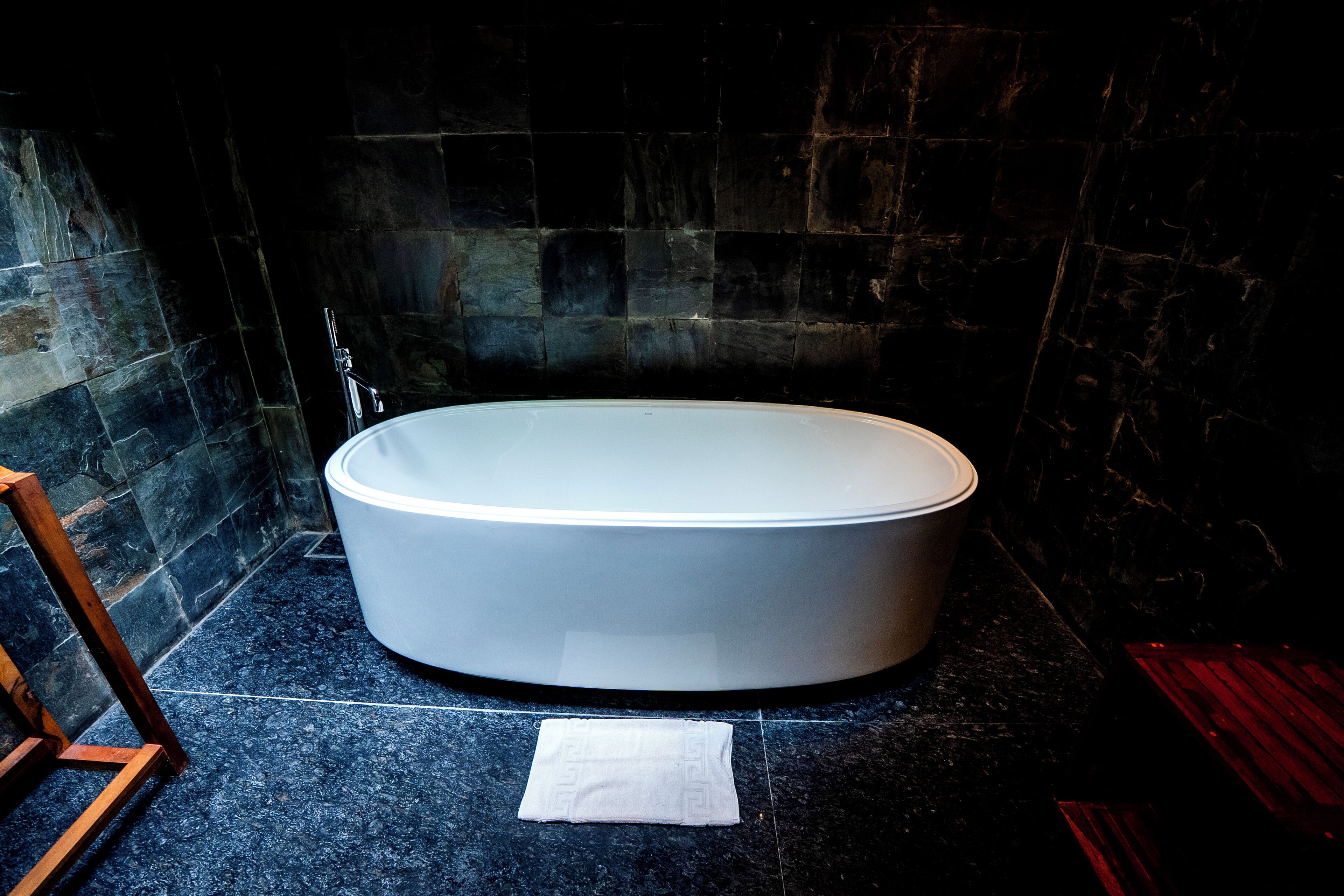 A modern bathroom with a freestanding bathtub, dark stone walls, and a wooden chair.