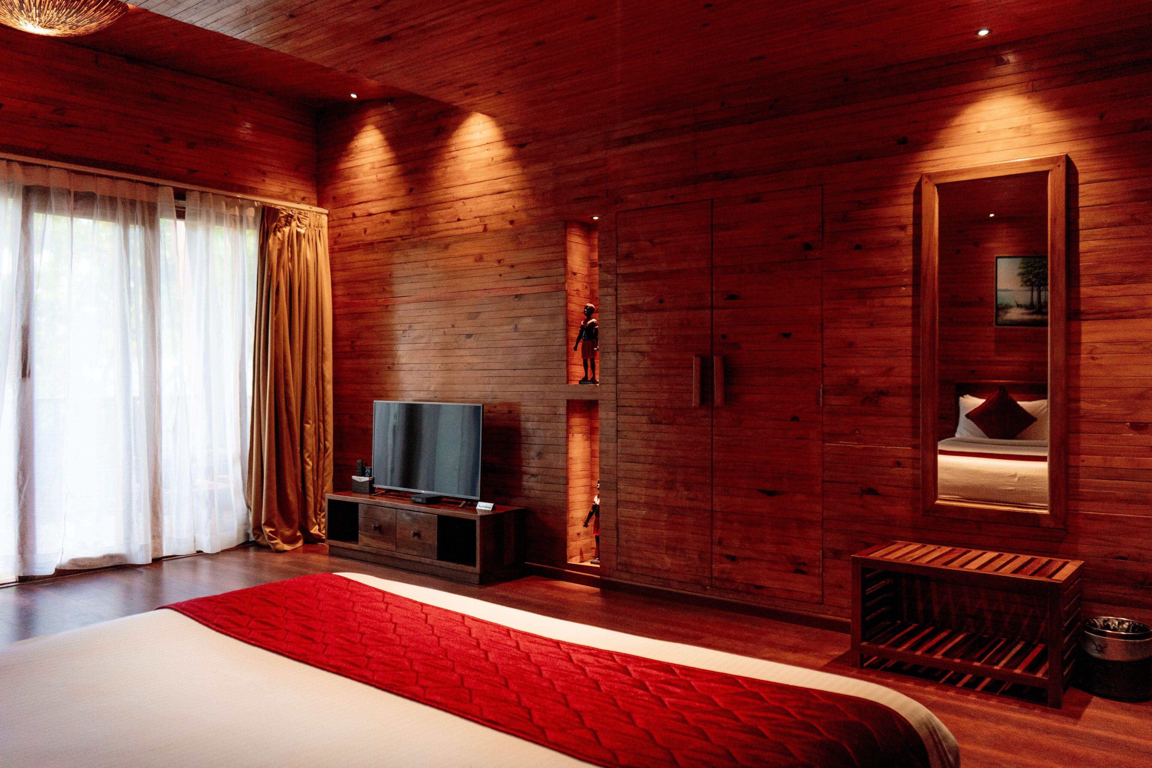 Una camera da letto moderna con pareti in legno, un letto grande con un tocco di colore rosso, una televisione su un supporto in legno e un piccolo sgabello di legno.