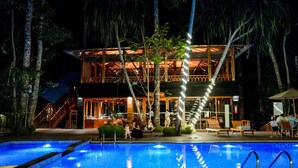 A pool area with a wooden deck, lounge chairs, and a building with a glass facade and wooden structure. - De Foret (Port Blair)