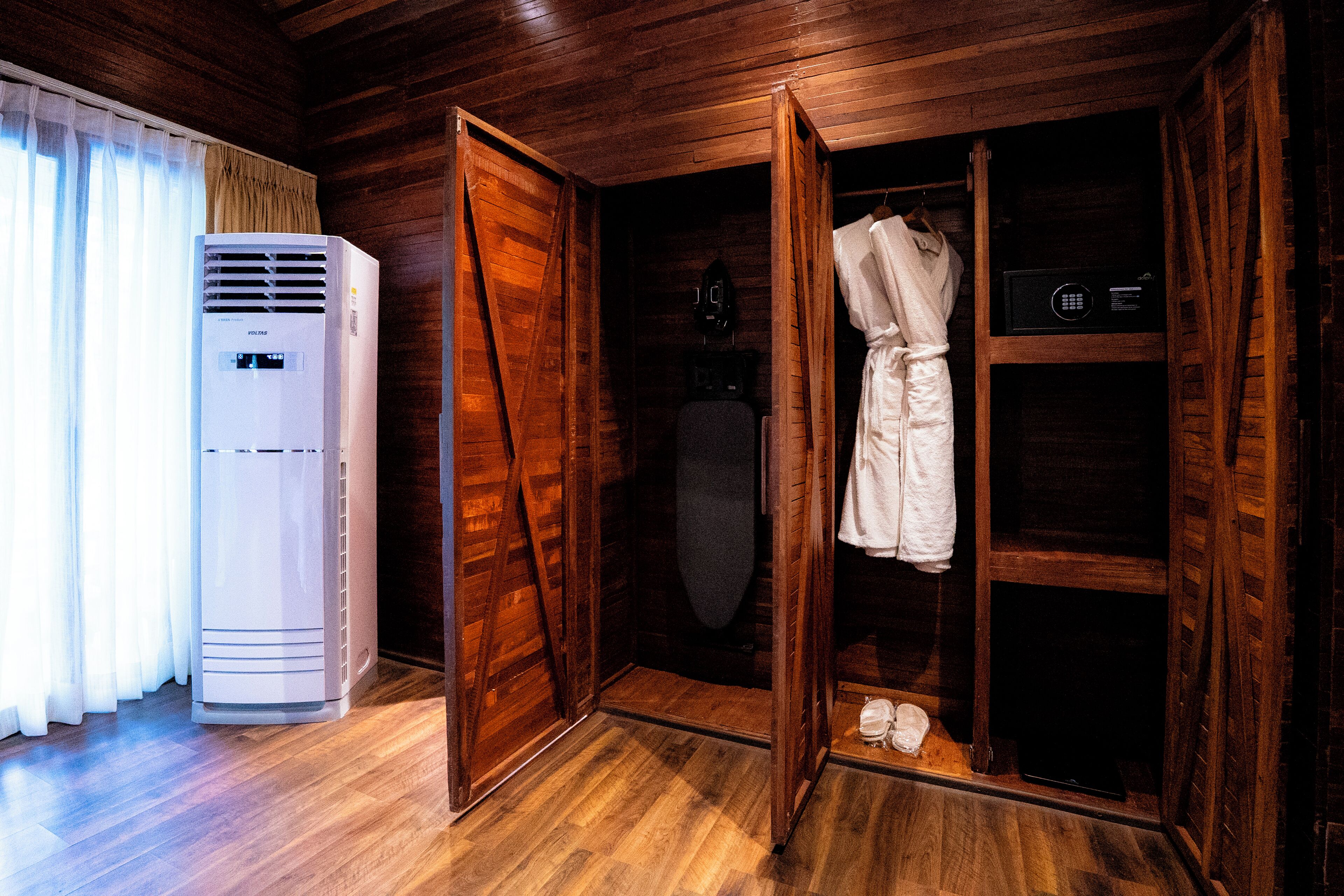 Una habitación de madera con un aire acondicionado blanco, un armario de madera con una bata colgando en su interior y un par de zapatos en el suelo.