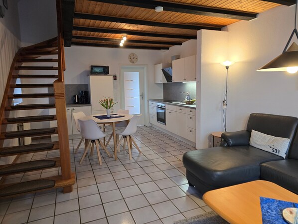 Eine moderne Küche mit weißen Schränken, ein Essbereich mit Tisch und Stühlen, ein schwarzes Ledersofa und eine Holztreppe.