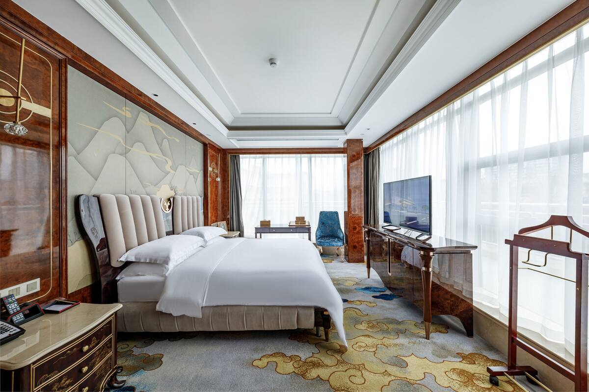 Quarto de hotel espaçoso com uma cama grande, uma mesa de escritório, televisão e vista para o exterior através de cortinas leves.