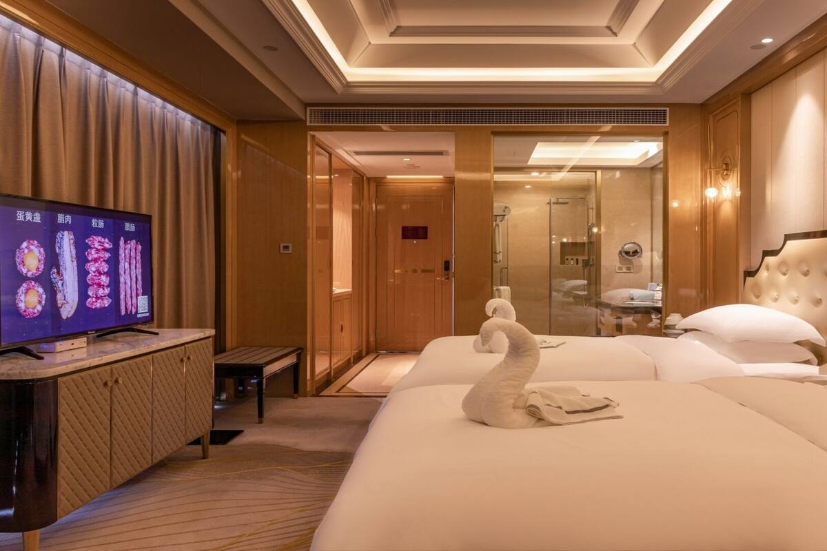 Quarto de hotel com uma cama grande, televisão sobre um console de madeira, área de chuveiro e porta para o banheiro.