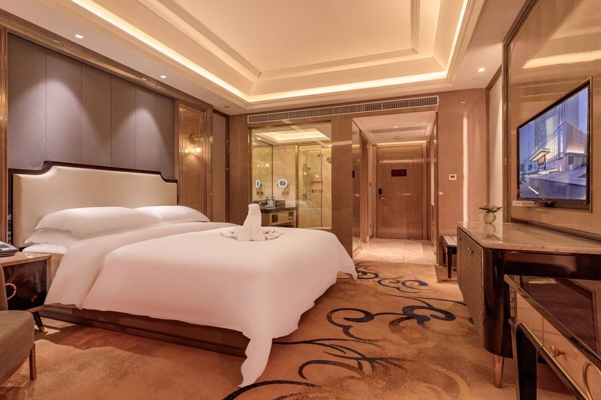 Quarto de hotel com uma cama grande, uma escrivaninha com telefone, televisão e banheiro com chuveiro de vidro.