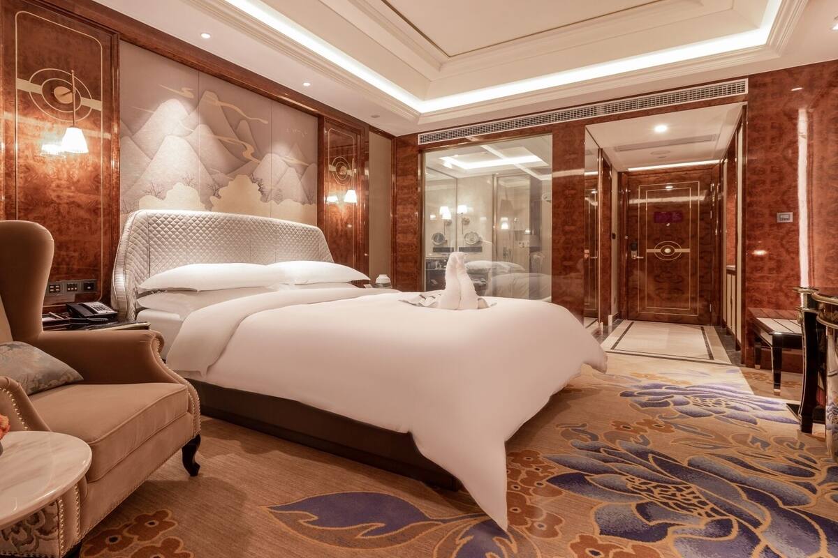 Quarto de hotel espaçoso com uma cama grande, área de estar com sofá e poltrona, pequena mesa lateral com um vaso de flores e um mural decorativo na parede.