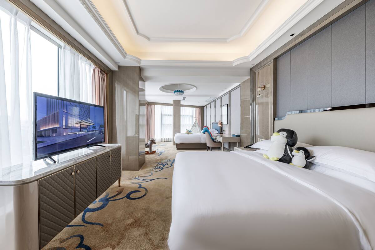 Quarto de hotel com uma cama grande, televisão sobre um suporte, escrivaninha com cadeira e janela com cortinas.
