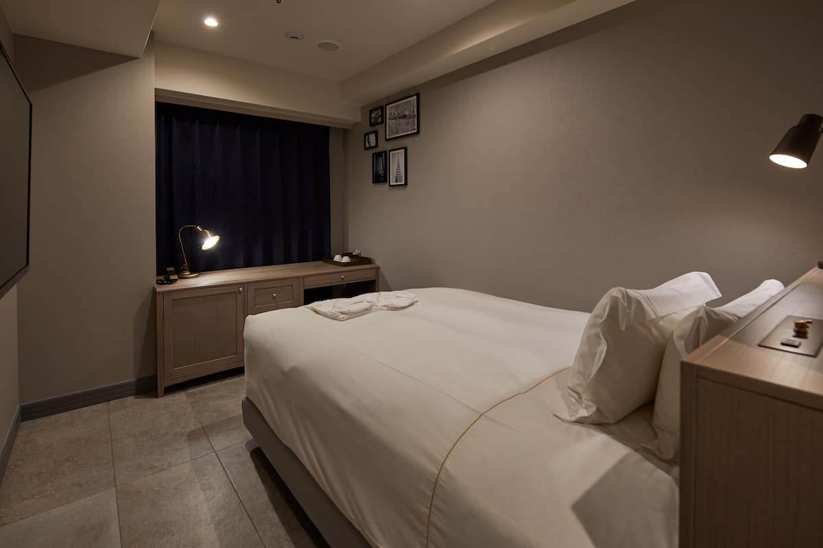 A hotel room with a large bed, a desk with a lamp, a wardrobe, and a window with curtains.