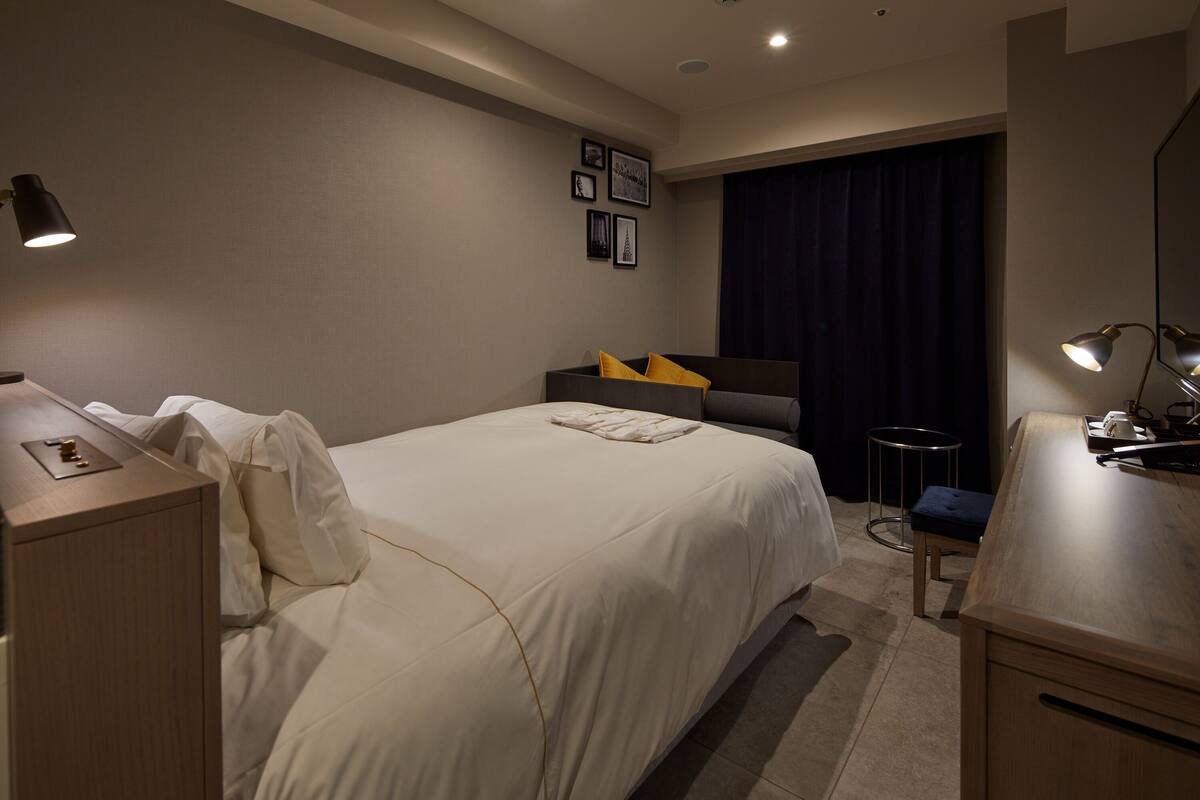 A hotel room with a bed, bedside table, lamp, and a small desk with a chair.