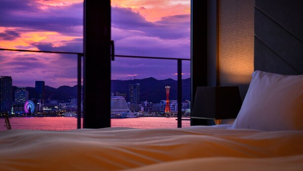 A hotel room with a bed, a nightstand, and a view of a cityscape at sunset. - Centurion Hotel & Spa Vintage Kobe (Kobe)