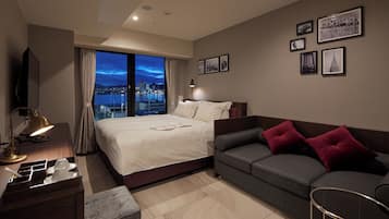 A hotel room with a large bed, a sofa, a desk with a lamp, and a city view through the window.