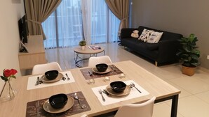 A modern living room with a dining area. There is a black sofa with decorative pillows, a dining table set with plates, bowls, and cutlery. A potted plant is on the floor next to the sofa.