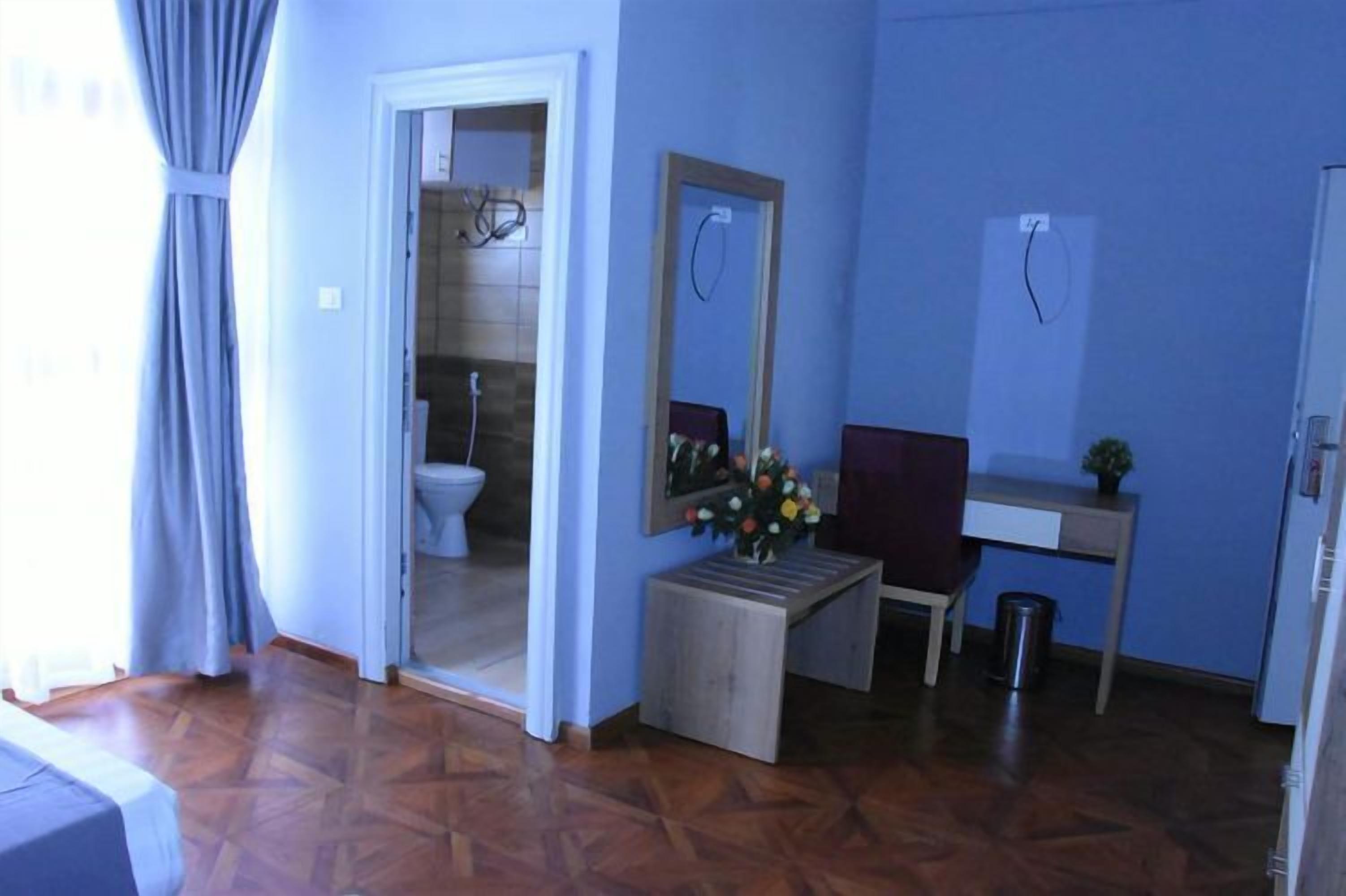 Una habitación con piso de parqué de madera, una cama, una mesita de madera con un florero y flores, un espejo, y una puerta que da a un baño.