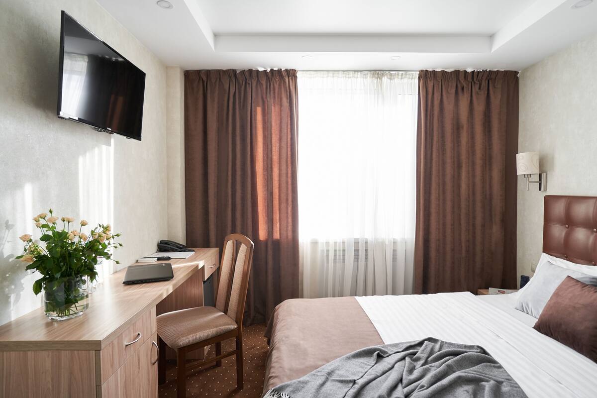 A hotel room with a bed, a desk with a laptop, a chair, a TV mounted on the wall, and curtains.