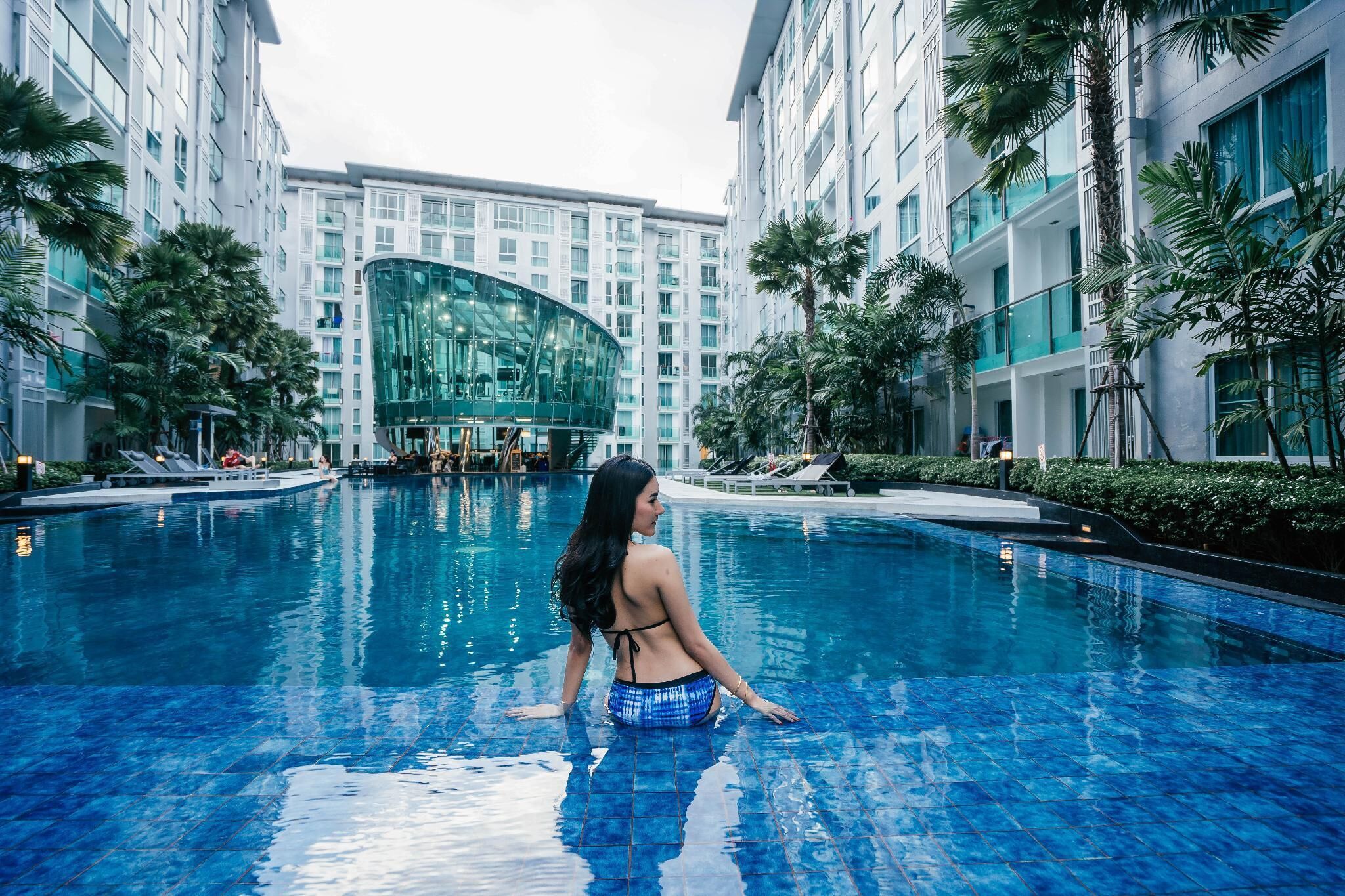 A swimming pool with a person sitting at the edge, surrounded by modern buildings and palm trees.