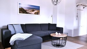 Ein modernes Wohnzimmer mit einer Eckcouch, einem runden Holztisch, einer Pendelleuchte und Strandbilder an der Wand.