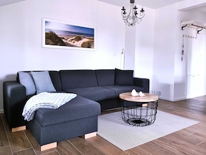 A modern living room with a sectional sofa, a round wooden coffee table, a chandelier, and a beach-themed wall art.