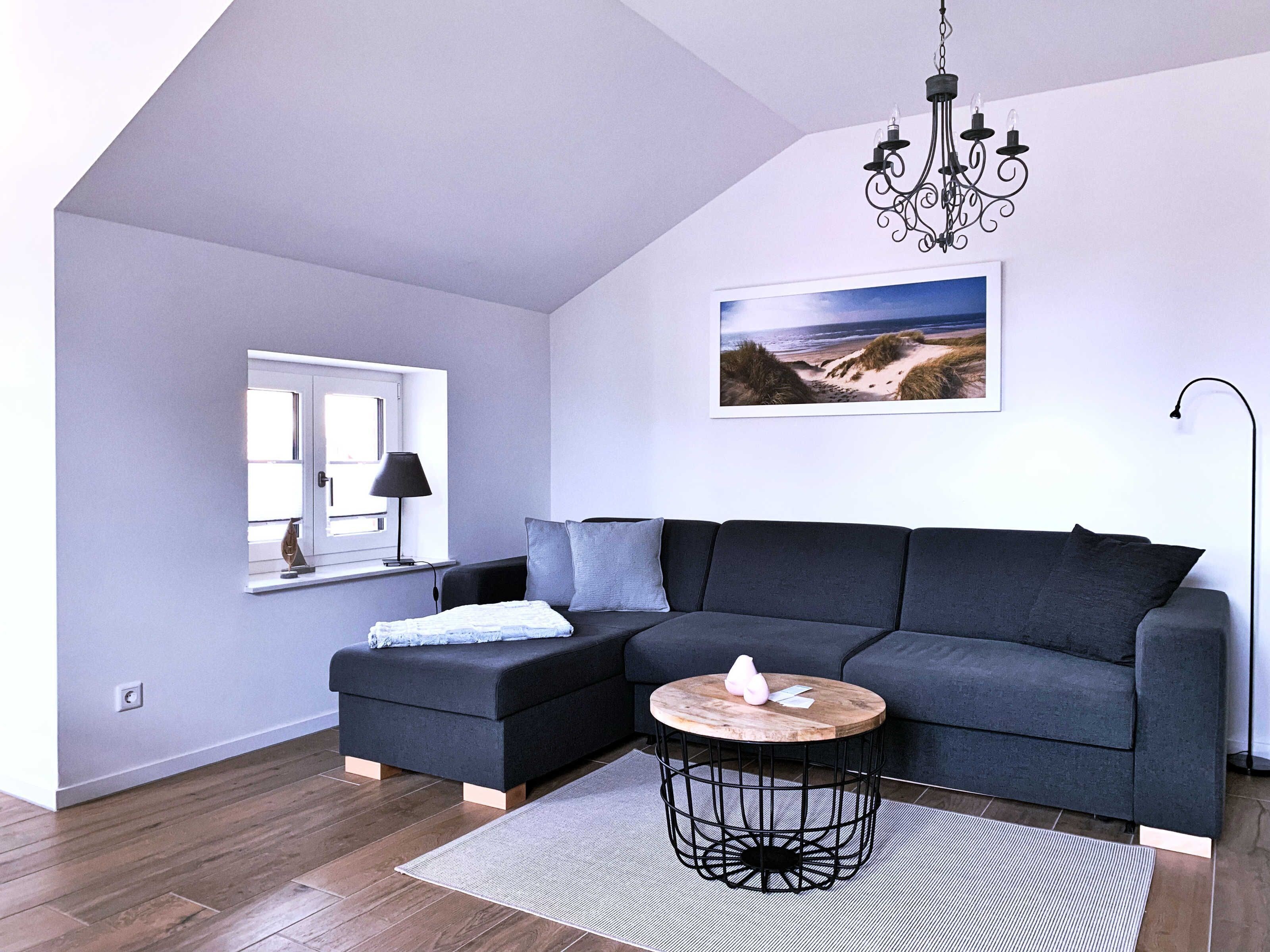 Una sala moderna con un sofá modular, una mesa de centro redonda, una araña, una ventana con vista a la playa y un cuadro en la pared.