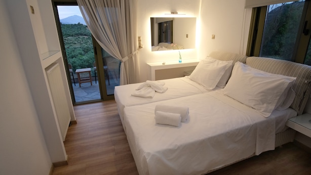 Uma cama de casal com roupa de cama e toalhas brancas, uma mesa de cabeceira com abajur, um espelho sobre a cama e vista para o verde através de uma porta de vidro deslizante.