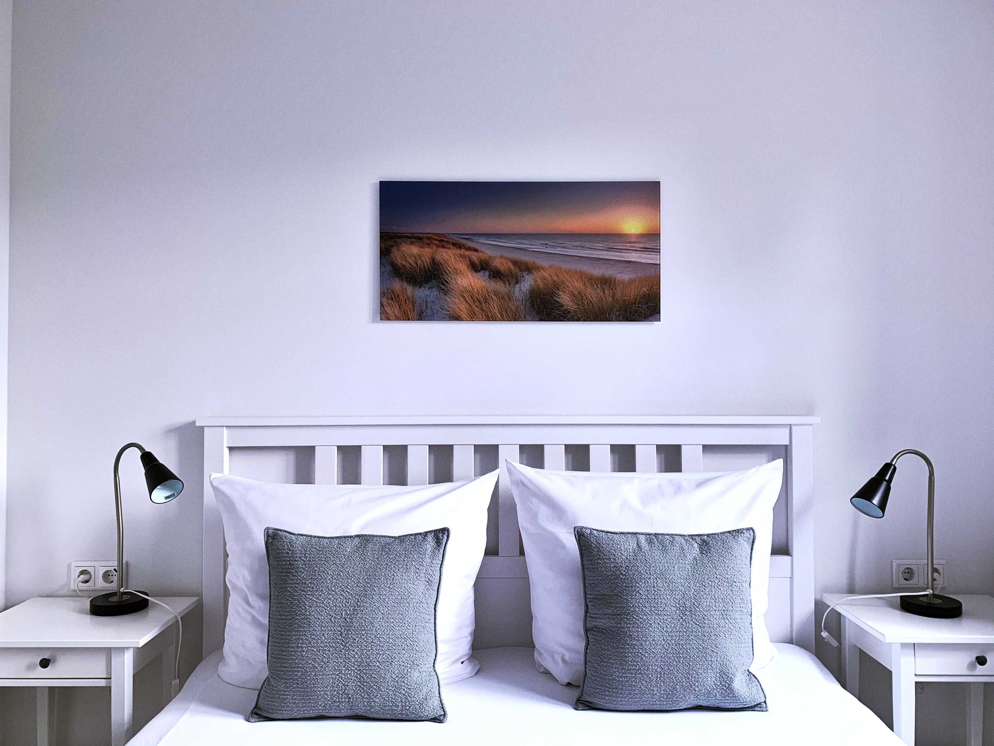 Een slaapkamer met een wit hoofdeinde, twee nachtkastjes met lampen, een wit bed met twee kussens, en een lijst met een afbeelding aan de muur.