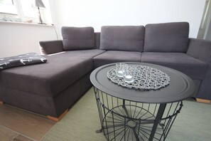 A grey sectional sofa with a matching ottoman, a round metal table with a woven coaster, and a green area rug.