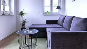 A modern living room with a grey sectional sofa, a round table with chairs, a potted plant, and a window with blinds. - FW Port Bridge, Germany (Wustrow)