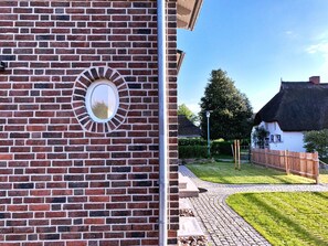 A brick wall with a circular window and a pathway leading to a thatched-roof building. - FW Backbord Peildeck (Wustrow)