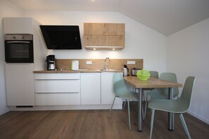 A modern kitchen with white cabinets, a built-in oven, a coffee machine, and a dining area with a wooden table and green chairs.