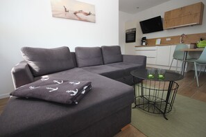 A modern living room with a grey sectional sofa, a small round table with two green glasses, a flat-screen TV mounted above a wooden kitchen counter, and a painting on the wall. - FW Backbord Peildeck (Wustrow)