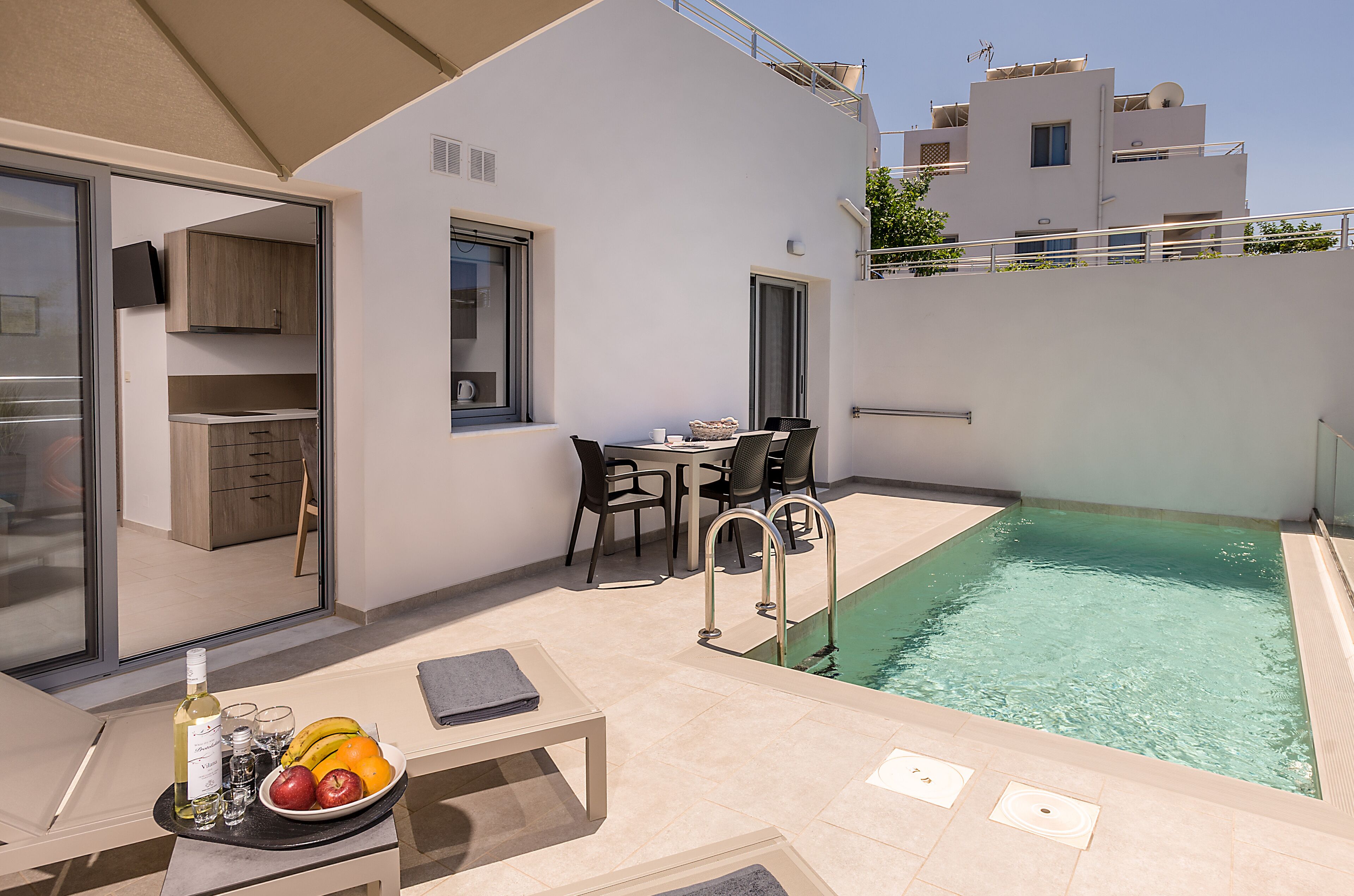 Un espace piscine extérieur moderne, doté d’un coin repas et d’une cuisine visible à travers une porte ouverte.