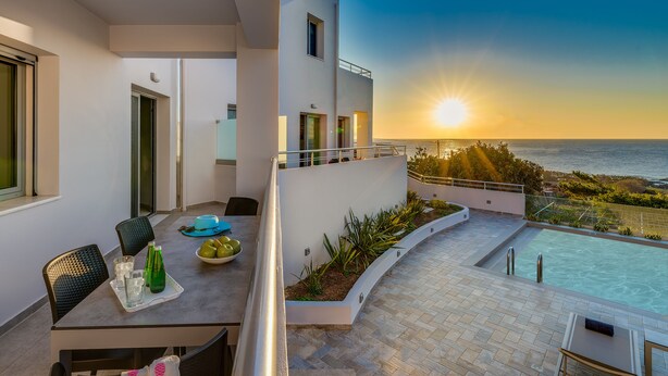 Un balcón moderno con piscina, zona de comedor y vistas a la puesta de sol sobre el mar.