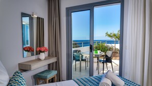 Habitación de hotel con cama, escritorio, silla, mesita, espejo y vista al mar a través de una puerta corrediza de vidrio.