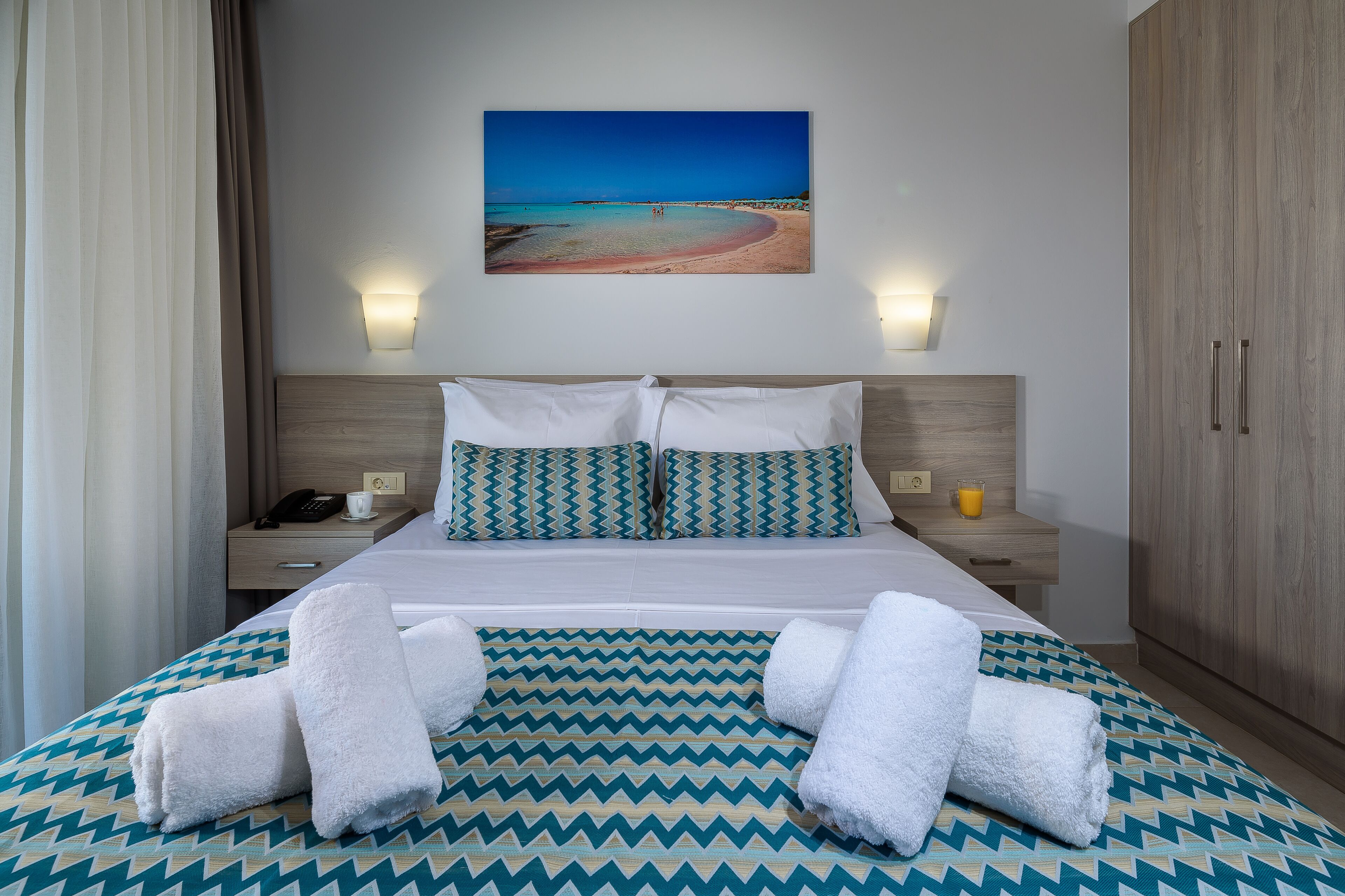Ein Hotelzimmer mit einem Bett, zwei Nachttischen, einem Wandlampenleuchter, einem Holzschrank und einem Gemälde mit Strandmotiv.