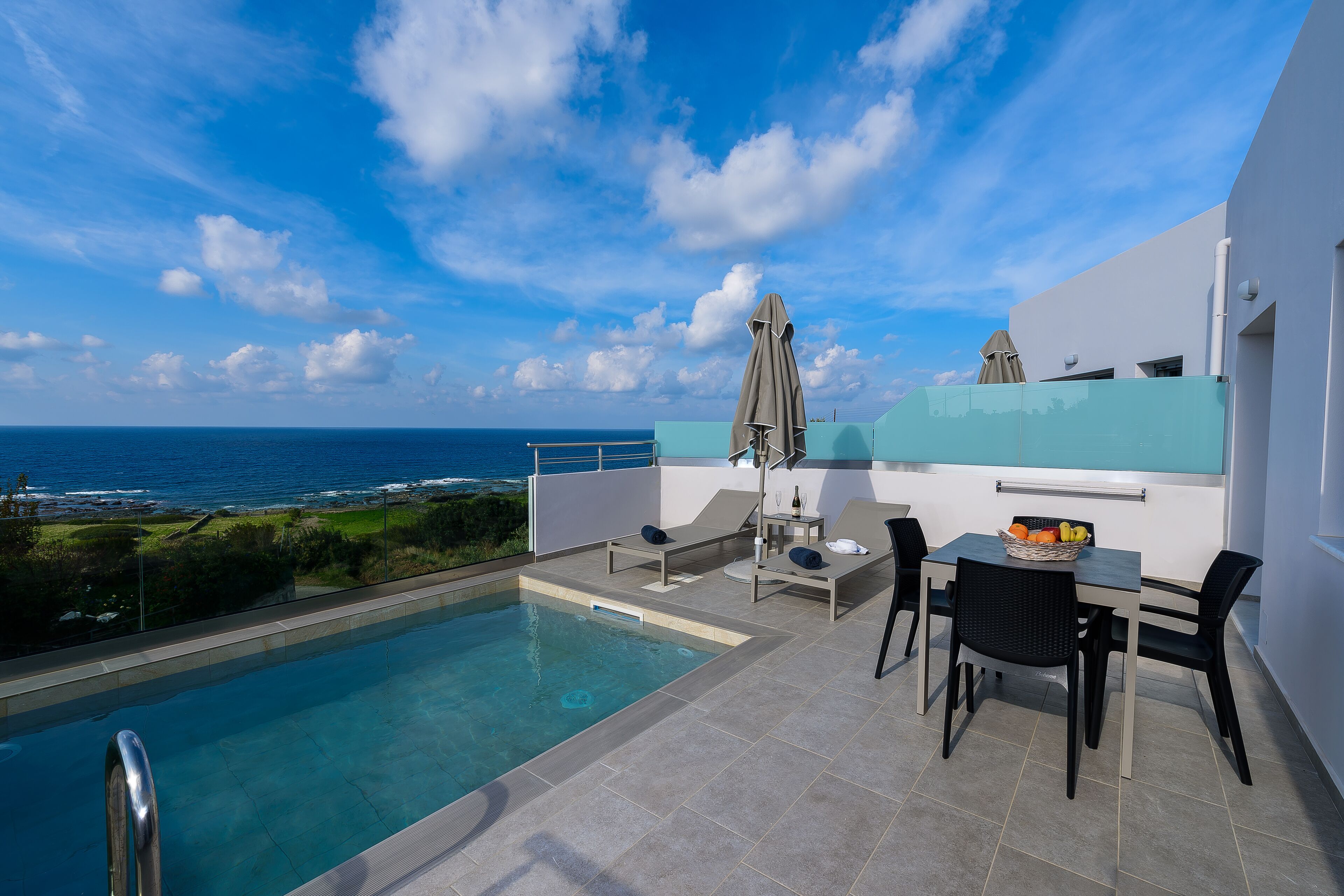 A rooftop pool area with a view of the ocean, a table with chairs, and a small table with a fruit bowl.