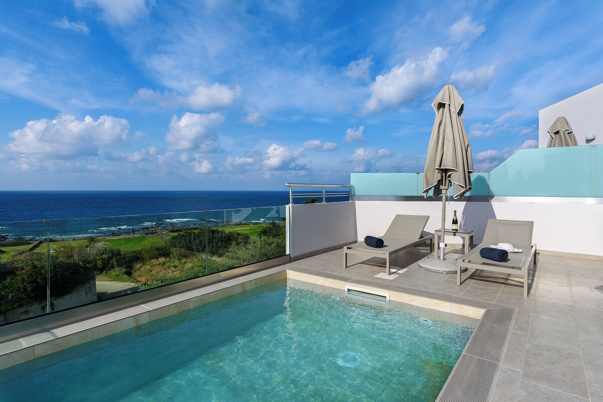 Une piscine sur le toit avec une eau d’un bleu limpide, deux chaises longues, une table avec une bouteille, et une vue sur l’océan.
