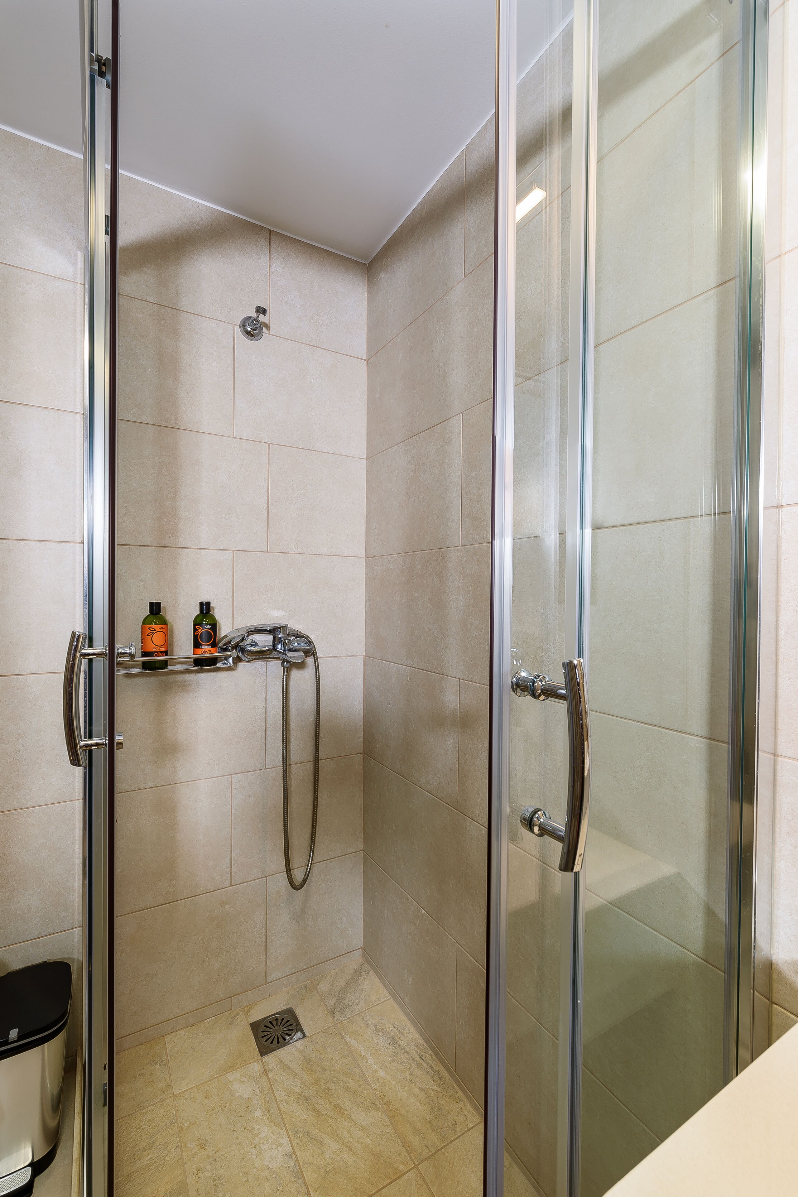A glass-enclosed shower stall with a handheld showerhead, a handheld shower attachment, and a shelf with toiletries.