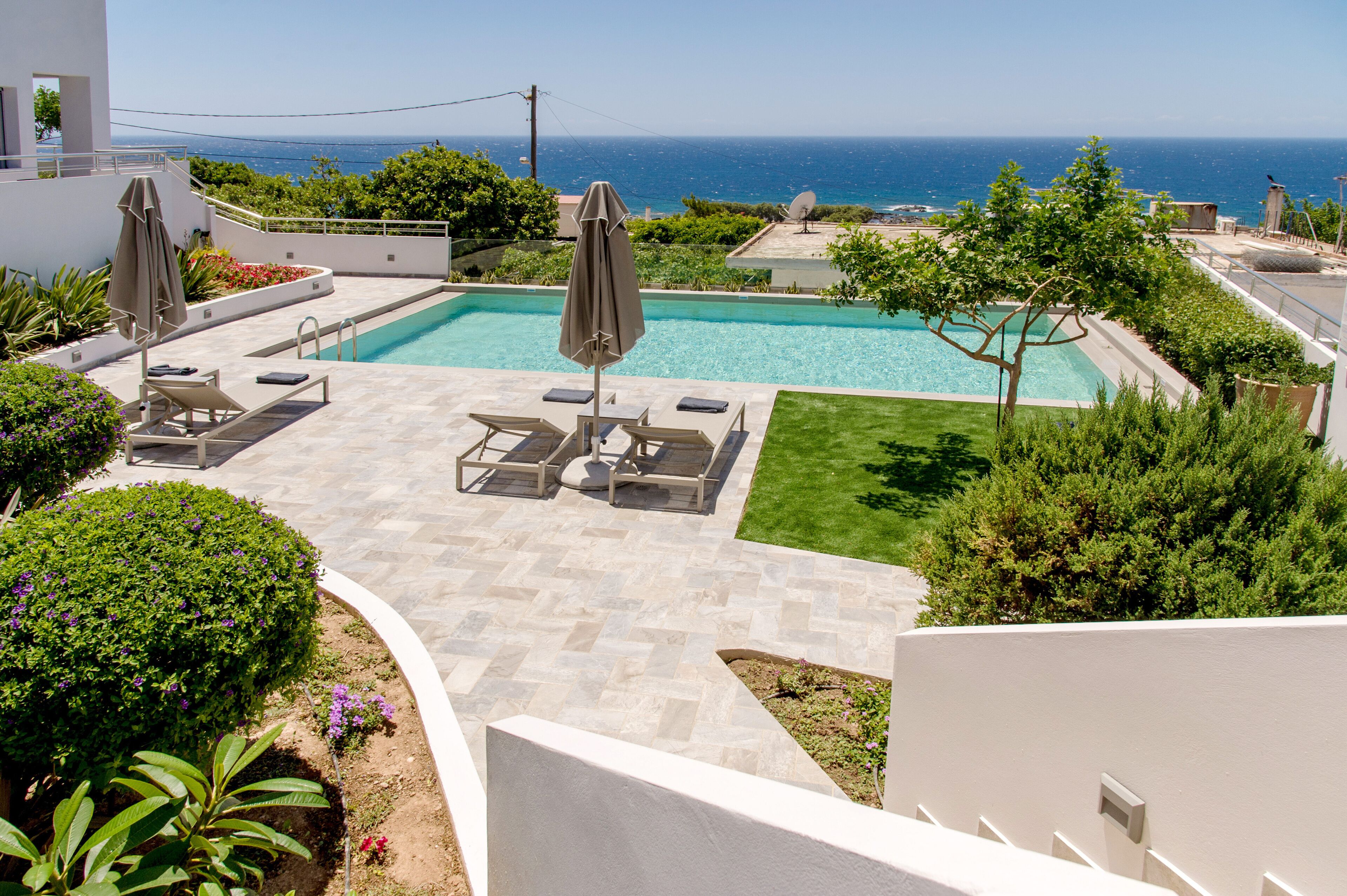 A pool area with lounge chairs and umbrellas, surrounded by greenery and a view of the sea.