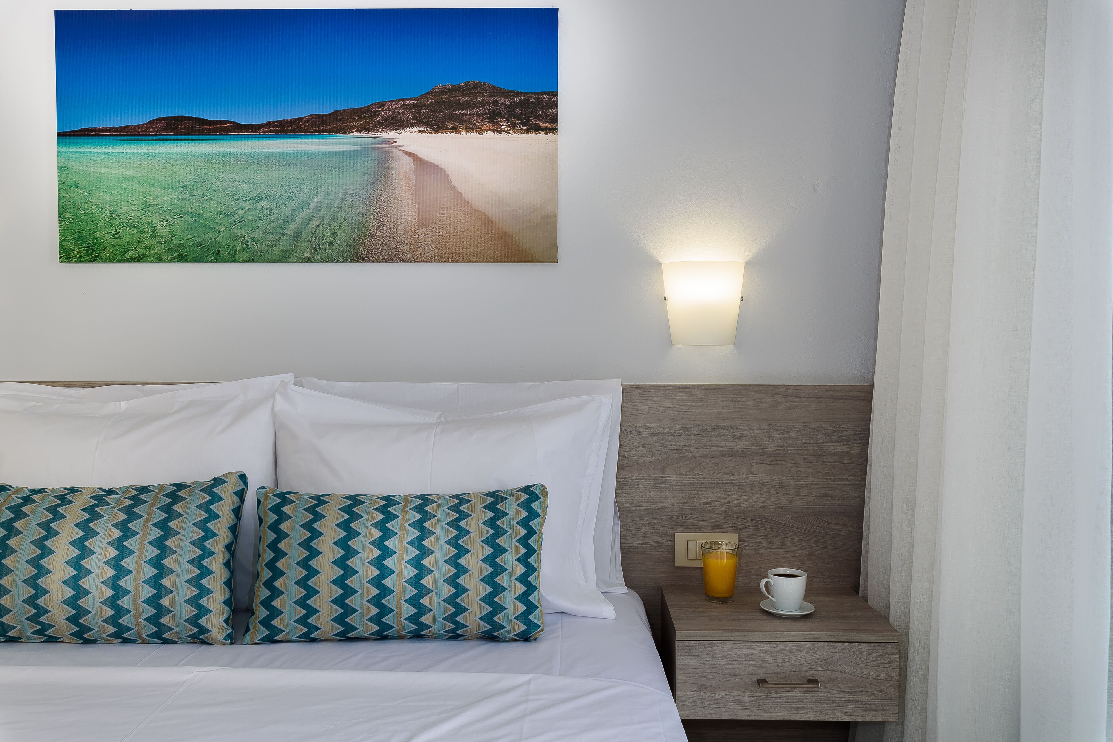 A hotel room with a bed, two patterned pillows, a nightstand with a cup of coffee, and a wall-mounted picture of a beach.