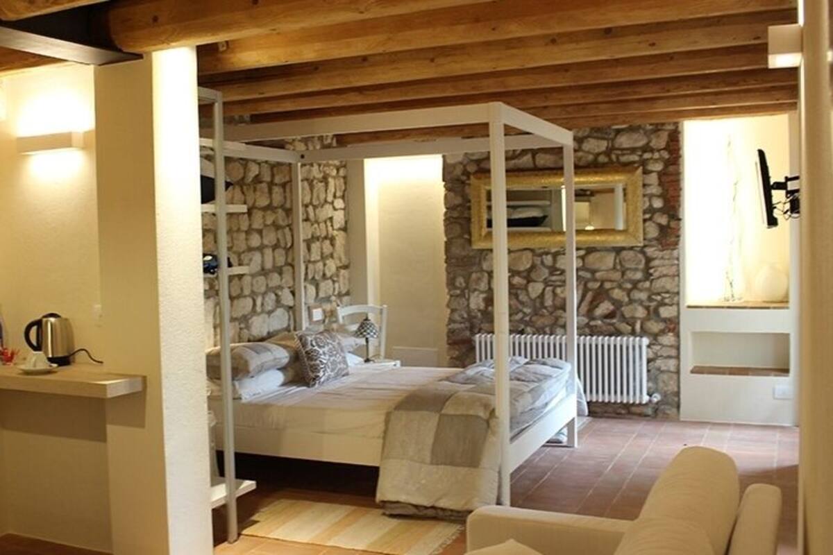 A bedroom with a stone wall, a bed with pillows, a bedside table with a kettle, and a chair.