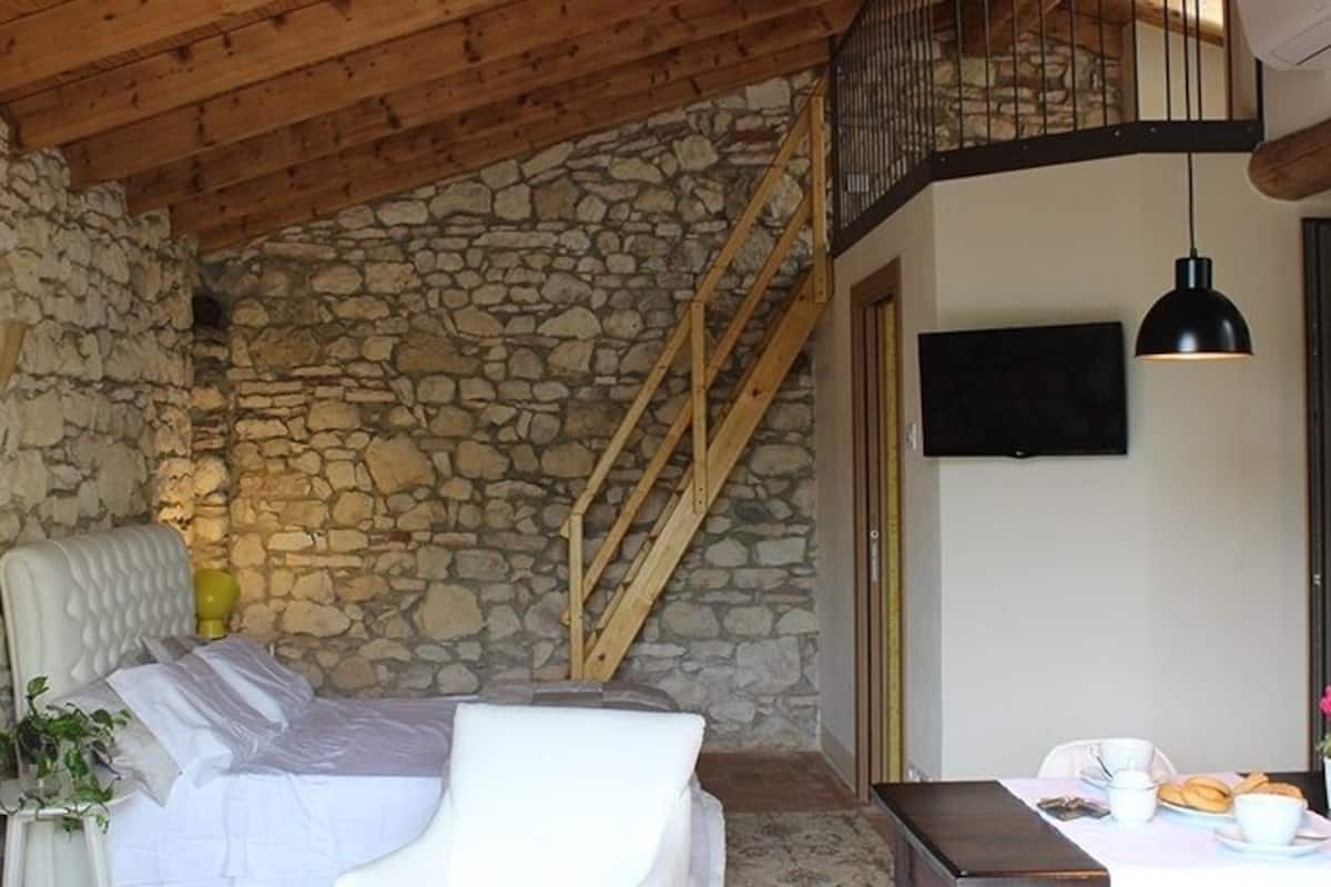 A cozy room with a stone wall, a bed, a wooden staircase, a TV, and a dining area with a table and chairs.