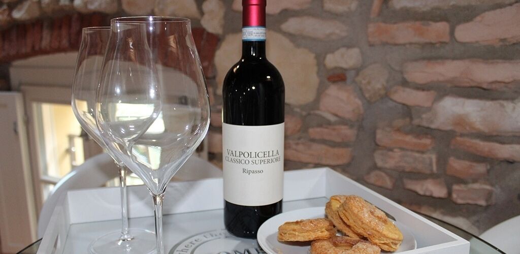 A bottle of Valpolicella Classico Superiore Ripasso wine, two empty wine glasses, and a plate with pastries on a table with a stone wall background.